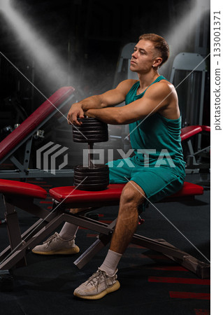 athletic person sits on a gym bench holding a dumbbell while taking a moment to rest. The setting is a fitness center filled with various gym equipment showcasing dedication to training. athletic person sits on a gym bench holding a dumbbell while taking a moment to rest. The setting is a fitness center filled with various gym equipment showcasing dedication to training. 133001371