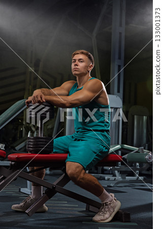 athletic individual sits on a weight bench in a gym taking a moment to rest while holding a set of dumbbells. The atmosphere is focused and intense as equipment surrounds them. 133001373