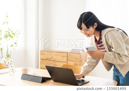 一位年輕女子一手拿著智慧型手機,正在檢查電腦。 一位年輕女子一手拿著智慧型手機,正在檢查電腦。 133001409