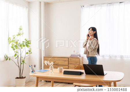 年輕女子一邊繞著桌子走動,一邊用智慧型手機通話。 年輕女子一邊繞著桌子走動,一邊用智慧型手機通話。 133001488