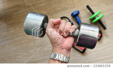 Close-up of a hand gripping a heavy dumbbell during a workout. Strength training and fitness exercise session at home or gym. Concept of determination, power, and healthy lifestyle. 133001559