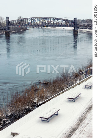 Snowy morning at Magdeburg Schleinufer shows calm river Elbe reflecting steel Hubbrucke bridge. Quiet winter path lined with blue lamps and benches creates serene urban landscape Snowy morning at Magdeburg Schleinufer shows calm river Elbe reflecting steel Hubbrucke bridge. Quiet winter path lined with blue lamps and benches creates serene urban landscape 133001850