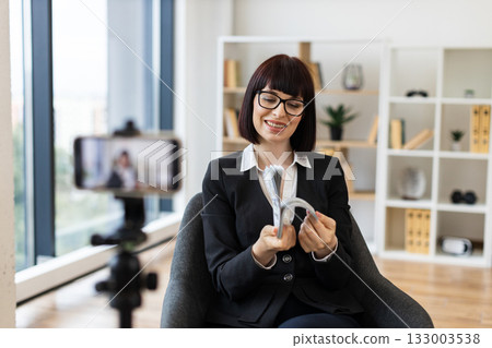 Professional setting with Caucasian adult woman presenting financial concepts through video blogging setup with cash. Professional setting with Caucasian adult woman presenting financial concepts through video blogging setup with cash. 133003538