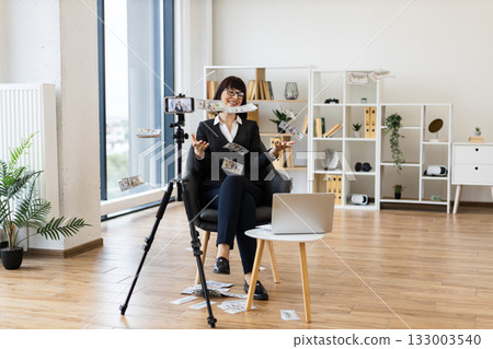 Caucasian woman in business attire sitting in modern office, blogging about investments on camera while confidently tossing dollar bills, showcasing entrepreneurial success, positivity. Caucasian woman in business attire sitting in modern office, blogging about investments on camera while confidently tossing dollar bills, showcasing entrepreneurial success, positivity. 133003540
