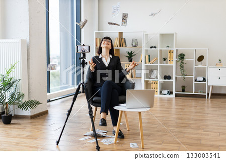 Caucasian woman in business attire sitting in modern office, blogging about investments on camera while confidently tossing dollar bills, showcasing entrepreneurial success, positivity. Caucasian woman in business attire sitting in modern office, blogging about investments on camera while confidently tossing dollar bills, showcasing entrepreneurial success, positivity. 133003541