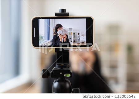 Caucasian woman in her thirties recording a financial vlog, dressed in a black business suit, holding dollar bills in hands, emphasizing wealth. Taken in a modern professional environment. Caucasian woman in her thirties recording a financial vlog, dressed in a black business suit, holding dollar bills in hands, emphasizing wealth. Taken in a modern professional environment. 133003542