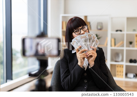 Caucasian woman in her thirties recording a financial vlog, dressed in a black business suit, holding dollar bills in hands, emphasizing wealth. Taken in a modern professional environment. Caucasian woman in her thirties recording a financial vlog, dressed in a black business suit, holding dollar bills in hands, emphasizing wealth. Taken in a modern professional environment. 133003543