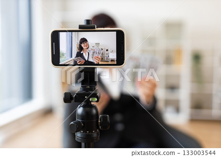 Caucasian young female professional blogging about financial tips, showing US dollar cash, seated in modern office with camera setup, wearing formal business attire, focusing on business communication Caucasian young female professional blogging about financial tips, showing US dollar cash, seated in modern office with camera setup, wearing formal business attire, focusing on business communication 133003544
