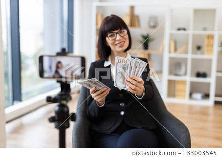 Caucasian young female professional blogging about financial tips, showing US dollar cash, seated in modern office with camera setup, wearing formal business attire, focusing on business communication Caucasian young female professional blogging about financial tips, showing US dollar cash, seated in modern office with camera setup, wearing formal business attire, focusing on business communication 133003545