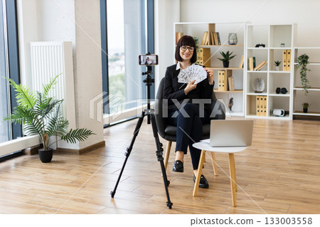 Caucasian woman, adult, wearing business attire, blogging on camera about investments holding cash dollars in modern office interior. Caucasian woman, adult, wearing business attire, blogging on camera about investments holding cash dollars in modern office interior. 133003558