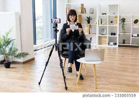 Caucasian woman in her 30s blogging on camera about financial topics holding cash dollars. Modern workspace background emphasizes professional context. Caucasian woman in her 30s blogging on camera about financial topics holding cash dollars. Modern workspace background emphasizes professional context. 133003559