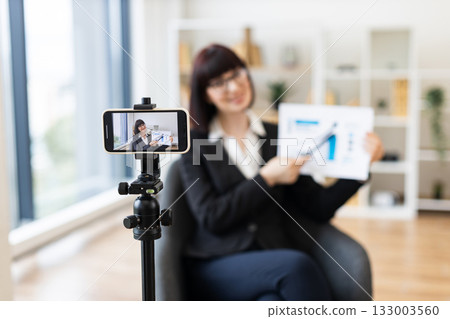 Caucasian woman in business suit recording video about revenue growth using graphs, modern office setup with natural light, professional presentation, concept communication and education. Caucasian woman in business suit recording video about revenue growth using graphs, modern office setup with natural light, professional presentation, concept communication and education. 133003560