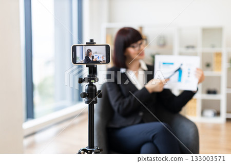 Caucasian woman in business suit recording video about revenue growth using graphs, modern office setup with natural light, professional presentation, concept communication and education. 133003571