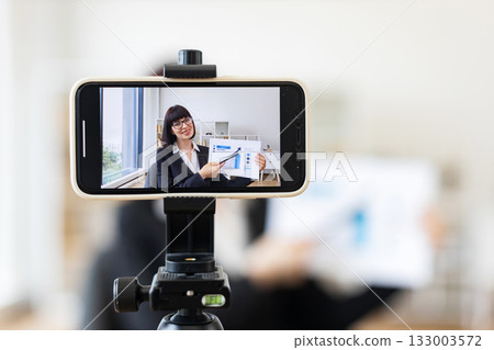 Caucasian woman presenting business charts and growth analysis while being recorded through smartphone setup Caucasian woman presenting business charts and growth analysis while being recorded through smartphone setup 133003572