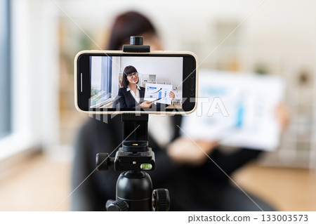 Caucasian woman presenting business charts and growth analysis while being recorded through smartphone setup Caucasian woman presenting business charts and growth analysis while being recorded through smartphone setup 133003573