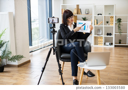 Caucasian woman presenting business analytics on camera through blogging using report and statistics chart. 133003580