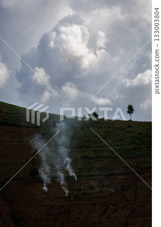 A photo of a lonely hill shows smoke rising from a small fire on a hillside farm under a dramatic cloud-filled sky. 133003804