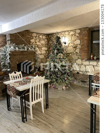 Christmas tree decorated with ornaments inside cozy rustic restaurant setting with empty chairs 133004178