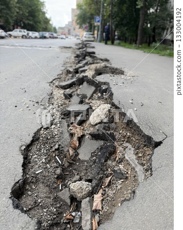 Damaged urban road with large crack showing broken asphalt and exposed dirt with leaves 133004192