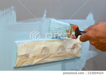 Painting the wall near an electrical outlet with a brush, utilizing masking tape to shield the outlet, during a home renovation task, focusing on neatness and interior maintenance. Painting the wall near an electrical outlet with a brush, utilizing masking tape to shield the outlet, during a home renovation task, focusing on neatness and interior maintenance. 133004424