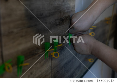 Hands carefully install wood-look ceramic wall tiles using leveling spacers. Tiles are laid using special levelers, ensuring precise and even alignment for a professional finish. 133004912