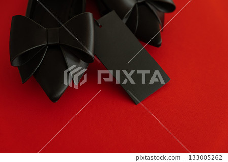 Elegant black pointed-toe shoes adorned with bows and a blank tag on red background Elegant black pointed-toe shoes adorned with bows and a blank tag on red background 133005262