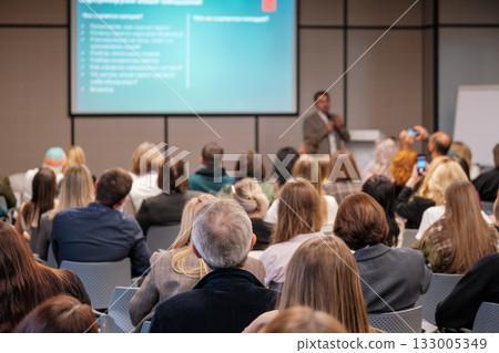 Engaged audience at a business conference listening to a speaker in a modern lecture hall with a large screen Engaged audience at a business conference listening to a speaker in a modern lecture hall with a large screen 133005349