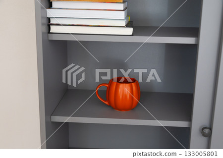 Orange mug on gray bookshelf under stack of books. Color accent, creativity, and cozy atmosphere of home office. 133005801