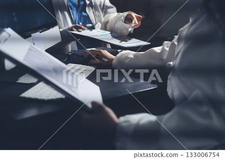 Two doctors and a female nurse meet at a table in the hospital, collaborating on medical tasks using laptops and computers 133006754