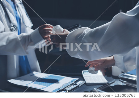Two doctors and a female nurse meet at a table in the hospital, collaborating on medical tasks using laptops and computers Two doctors and a female nurse meet at a table in the hospital, collaborating on medical tasks using laptops and computers 133006765