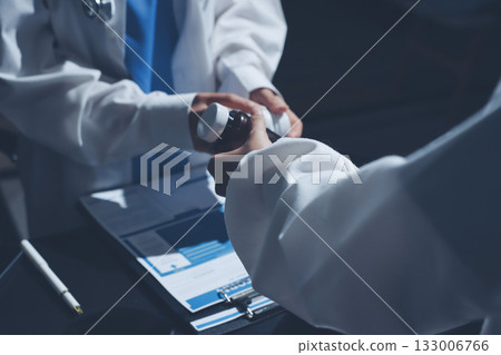Two doctors and a female nurse meet at a table in the hospital, collaborating on medical tasks using laptops and computers Two doctors and a female nurse meet at a table in the hospital, collaborating on medical tasks using laptops and computers 133006766