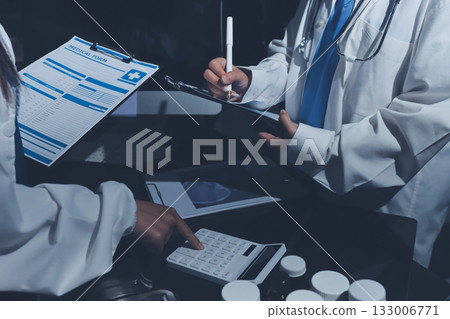 Two doctors and a female nurse meet at a table in the hospital, collaborating on medical tasks using laptops and computers Two doctors and a female nurse meet at a table in the hospital, collaborating on medical tasks using laptops and computers 133006771