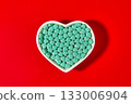 Turquoise colored cardiology pills are poured into a heart-shaped bowl on a red background. 133006904