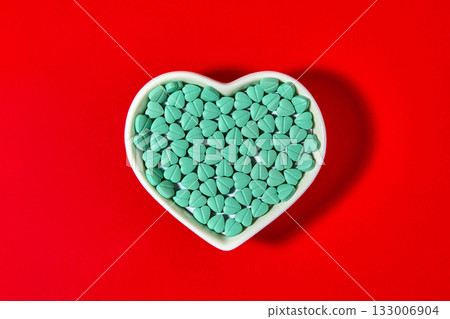Turquoise colored cardiology pills are poured into a heart-shaped bowl on a red background. Turquoise colored cardiology pills are poured into a heart-shaped bowl on a red background. 133006904