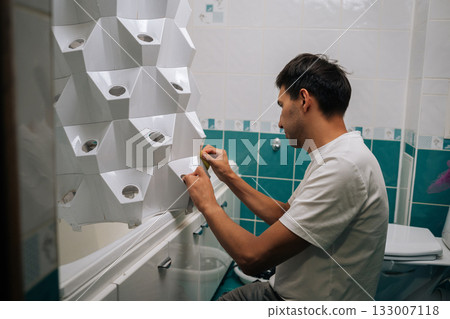 Gardener male wiping vertical hydroponic tower garden with yellow sponge, maintaining clean growing environment in contemporary bathroom space. Concept of healthy and sustainable cultivation. 133007118