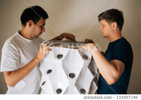 Agricultural technicians assembling vertical hydroponic tower garden indoors, carefully placing top part with grow light onto the structure, ready to start growing fresh produce at home. 133007146