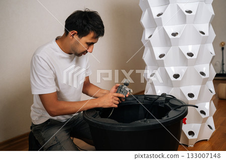 Focused home gardener installing water pump in reservoir of aeroponic vertical farming system, cultivating plants without soil in innovative and sustainable way indoors. Concept of urban farming. 133007148