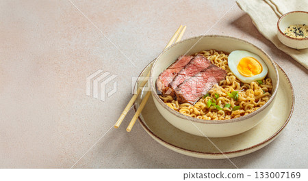 Ramen or asian style noodles soup with beef, egg and green onions in bowl on light beige background, space for text 133007169