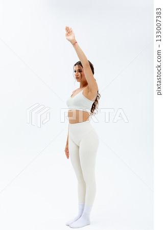Fitness enthusiast in white sportswear raises arm confidently upwards, Female athlete in bright activewear stretches on studio floor with dedicated focus and energy 133007383