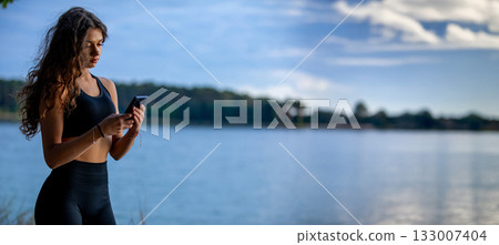 A Young Woman is Relaxing by the Calm Water While Joyfully Using Her Smartphone A Young Woman is Relaxing by the Calm Water While Joyfully Using Her Smartphone 133007404