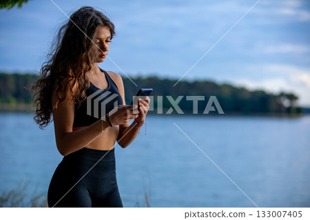 A young woman in active wear checks her smartphone by the water, enjoying the calm A young woman in active wear checks her smartphone by the water, enjoying the calm 133007405