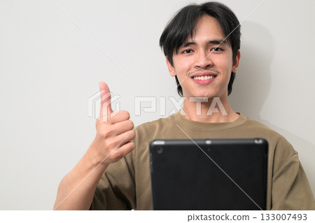 Portrait of happy young Asian Thai man using tablet computer against white wall giving thumbs up Portrait of happy young Asian Thai man using tablet computer against white wall giving thumbs up 133007493