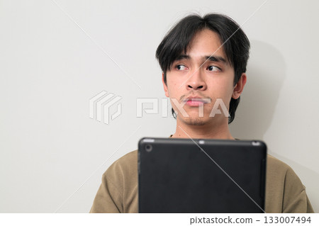 Portrait of handsome Young Asian Thai man using digital tablet computer against white wall thinking Portrait of handsome Young Asian Thai man using digital tablet computer against white wall thinking 133007494