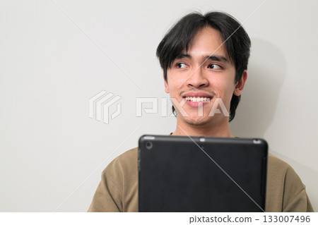 Portrait of happy young Asian Thai man using digital tablet computer while thinking Portrait of happy young Asian Thai man using digital tablet computer while thinking 133007496