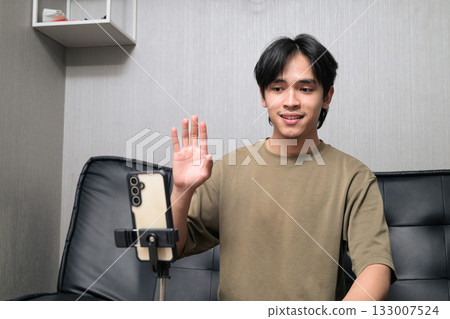 Portrait of handsome Young Asian Thai man vlogging with phone while sitting on sofa at home Portrait of handsome Young Asian Thai man vlogging with phone while sitting on sofa at home 133007524