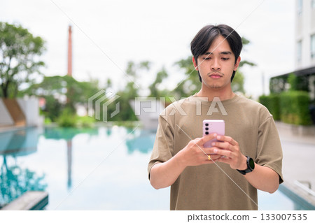 Young Asian Thai man in early 20s relaxing next to swimming pool on summer using phone 133007535