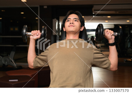 Portrait of fit young Asian Thai man in fitness gym at night doing shoulder press with dumbbells 133007570