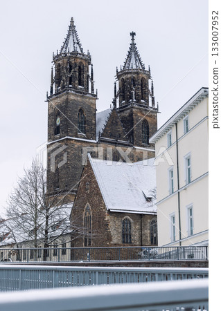 Snow-covered Magdeburg Cathedral towers rise against pale winter sky framed by modern building. Gothic architecture and frosty details create serene contrast of history and contemporary city scene 133007952
