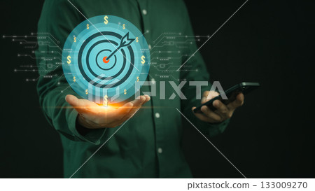 Businessman holding a smartphone with a glowing holographic target and dollar signs, symbolizing business goals, financial success, ROI, digital marketing, investment strategy and revenue growth. Businessman holding a smartphone with a glowing holographic target and dollar signs, symbolizing business goals, financial success, ROI, digital marketing, investment strategy and revenue growth. 133009270
