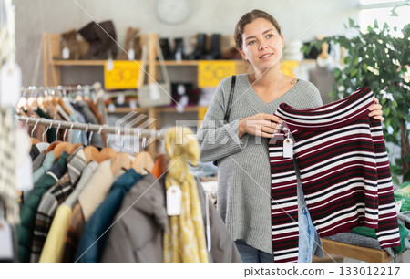 Interested positive pregnant european young woman shopping new sweaters in store. Sale of winter clothing collection 133012217